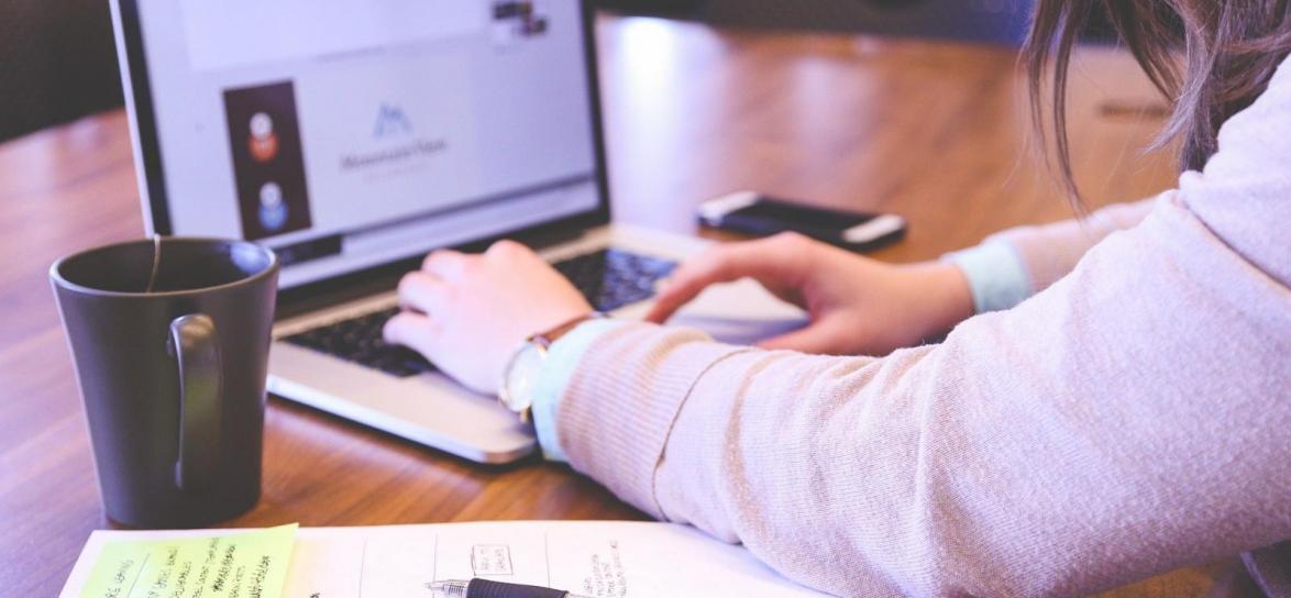 Osoba pisząca na klawiaturze laptopa, obok kawa i dokumenty